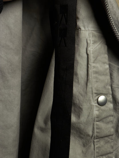 Close-up detail of a jacket showing a light gray fabric exterior, a black vertical inner panel with tonal logo stitching, a silver snap button, and a zipper.
