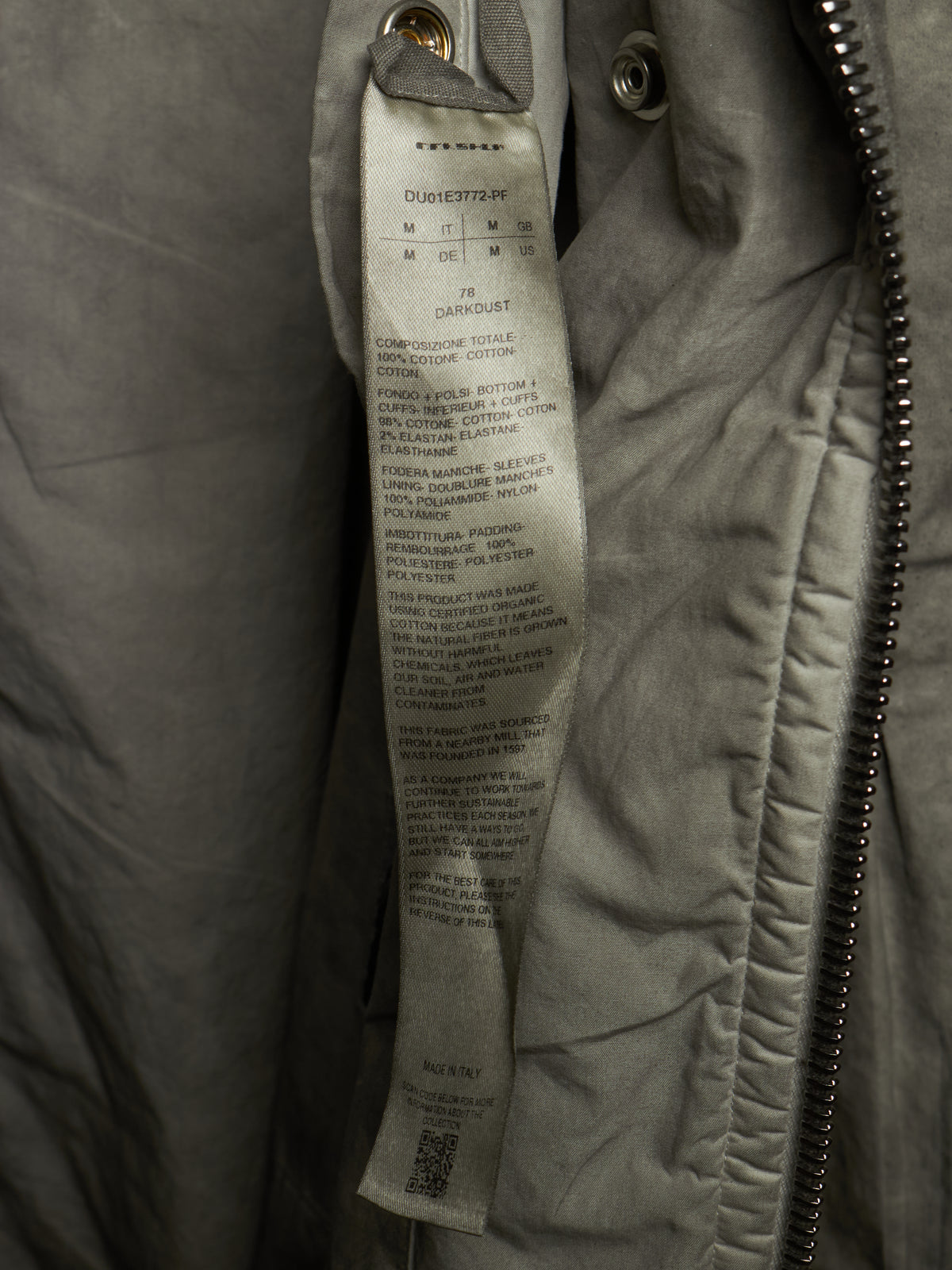 Close-up of a gray quilted jacket with a sewn-in size and care label visible, next to a metal zipper.