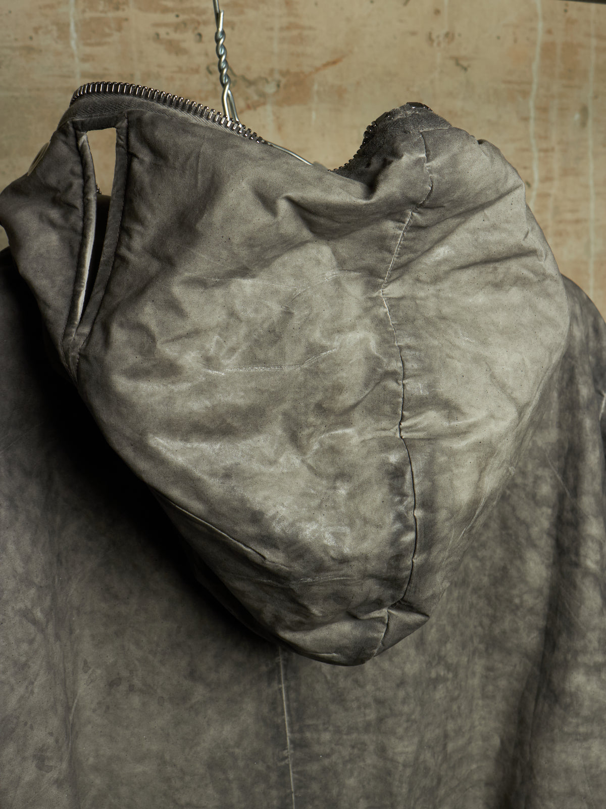 A close-up of a dark gray hooded jacket or coat hanging against a textured beige wall, showing the hood, zipper, and fabric texture.