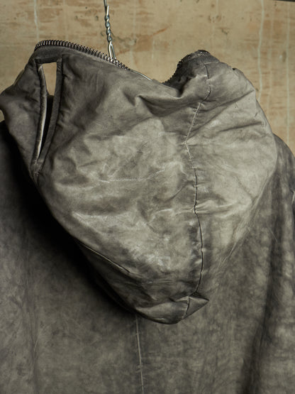 A close-up of a dark gray hooded jacket or coat hanging against a textured beige wall, showing the hood, zipper, and fabric texture.