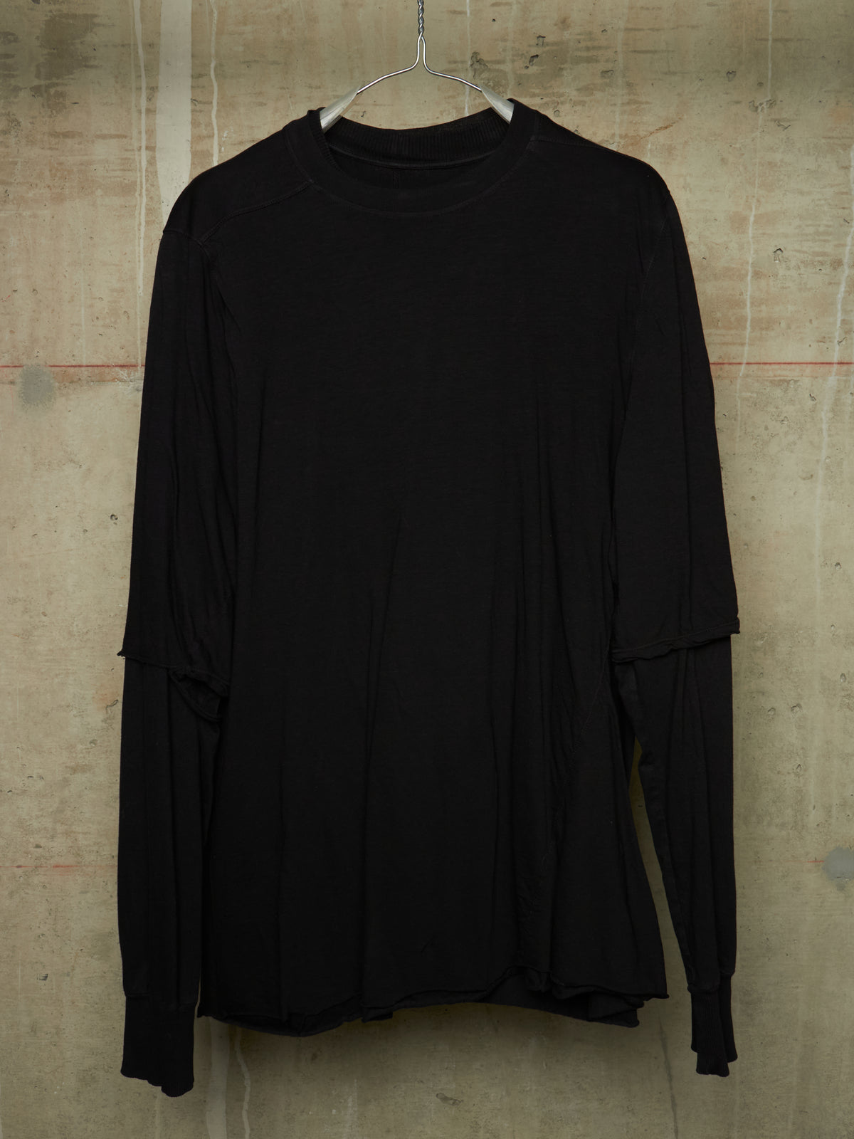 A black long-sleeve top with layered, asymmetrical sleeves hanging on a metal hanger against a textured concrete wall.
