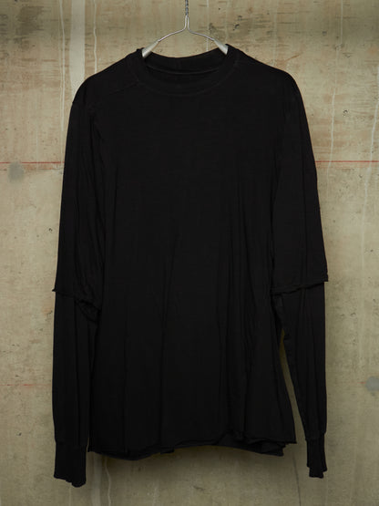 A black long-sleeve top with layered, asymmetrical sleeves hanging on a metal hanger against a textured concrete wall.
