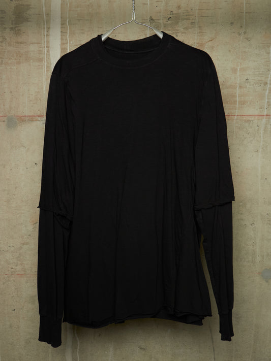 A black long-sleeve top with layered, asymmetrical sleeves hanging on a metal hanger against a textured concrete wall.
