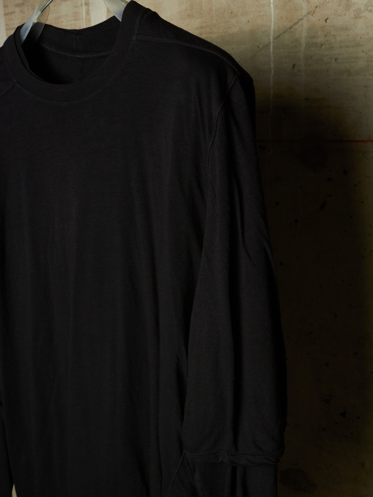 A black long-sleeve crewneck shirt hanging on a hanger against a textured concrete wall.