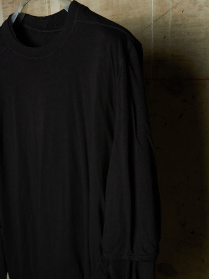 A black long-sleeve crewneck shirt hanging on a hanger against a textured concrete wall.