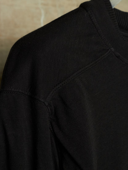 Close-up detail of a black knit crewneck sweater, showing the shoulder seam, ribbed collar, and fine fabric texture.
