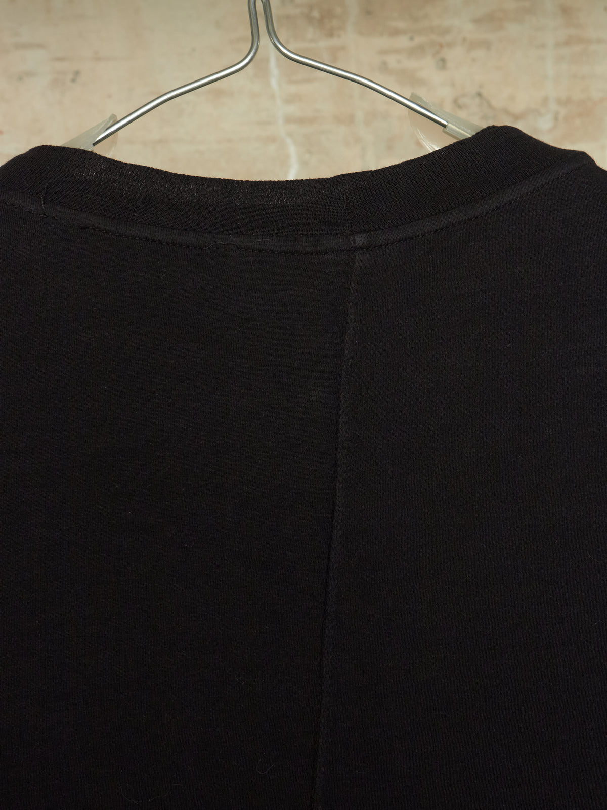 Close-up of the back of a black crewneck sweatshirt hanging on a metal hanger against a light beige textured wall.