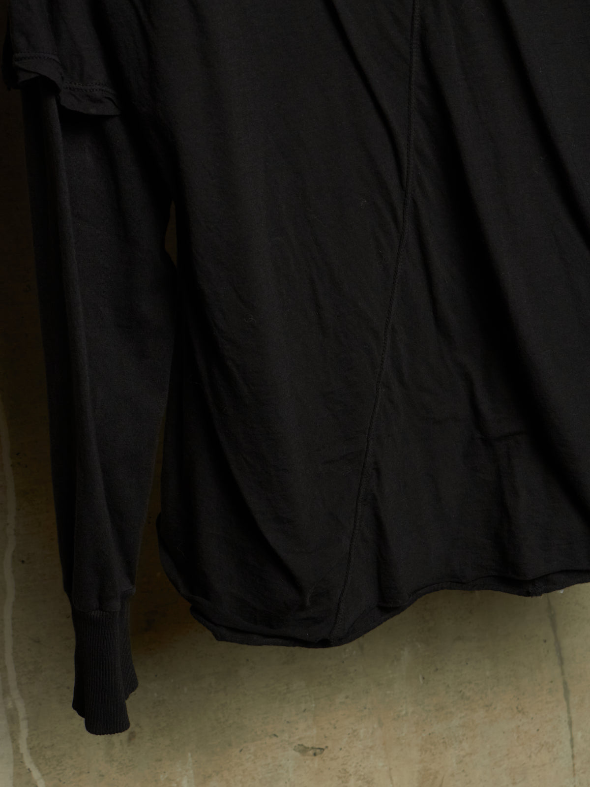 A close-up of a black long-sleeve top or dress with gathered ruffle detailing along the shoulder, hanging against a neutral concrete wall.
