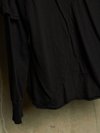 A close-up of a black long-sleeve top or dress with gathered ruffle detailing along the shoulder, hanging against a neutral concrete wall.