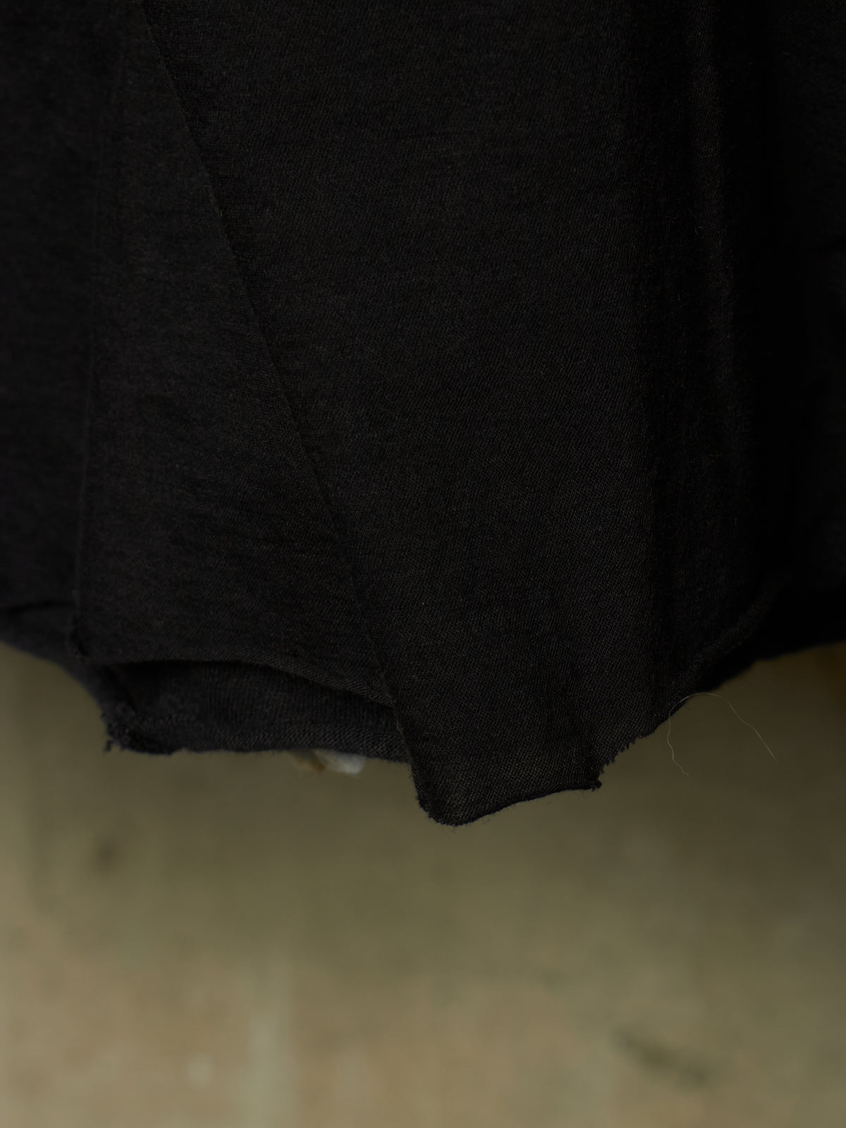 A close-up of a black fabric garment, showing the texture and hem of the material against a neutral beige background.
