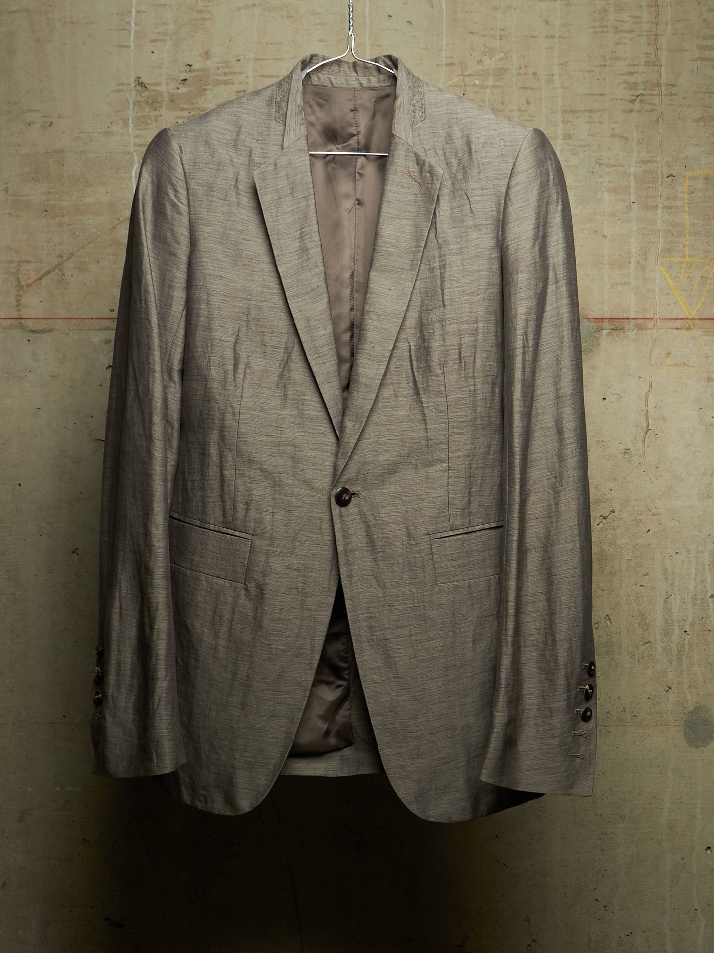 A single-breasted blazer jacket in a textured taupe-gray fabric, hanging on a metal hanger against a distressed concrete wall.