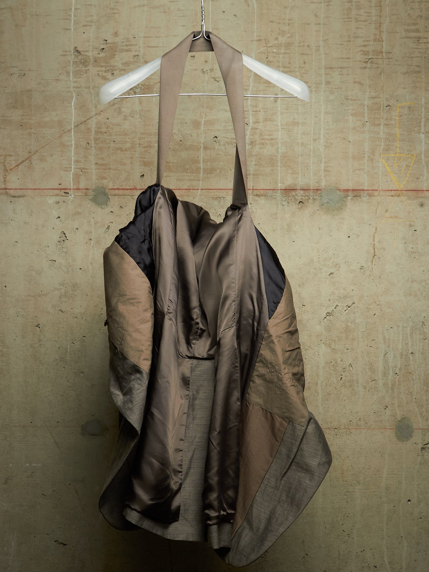 A draped, asymmetrical garment or dress made from layered taupe, brown, and black fabrics, hanging from a white plastic hanger against a textured concrete wall.
