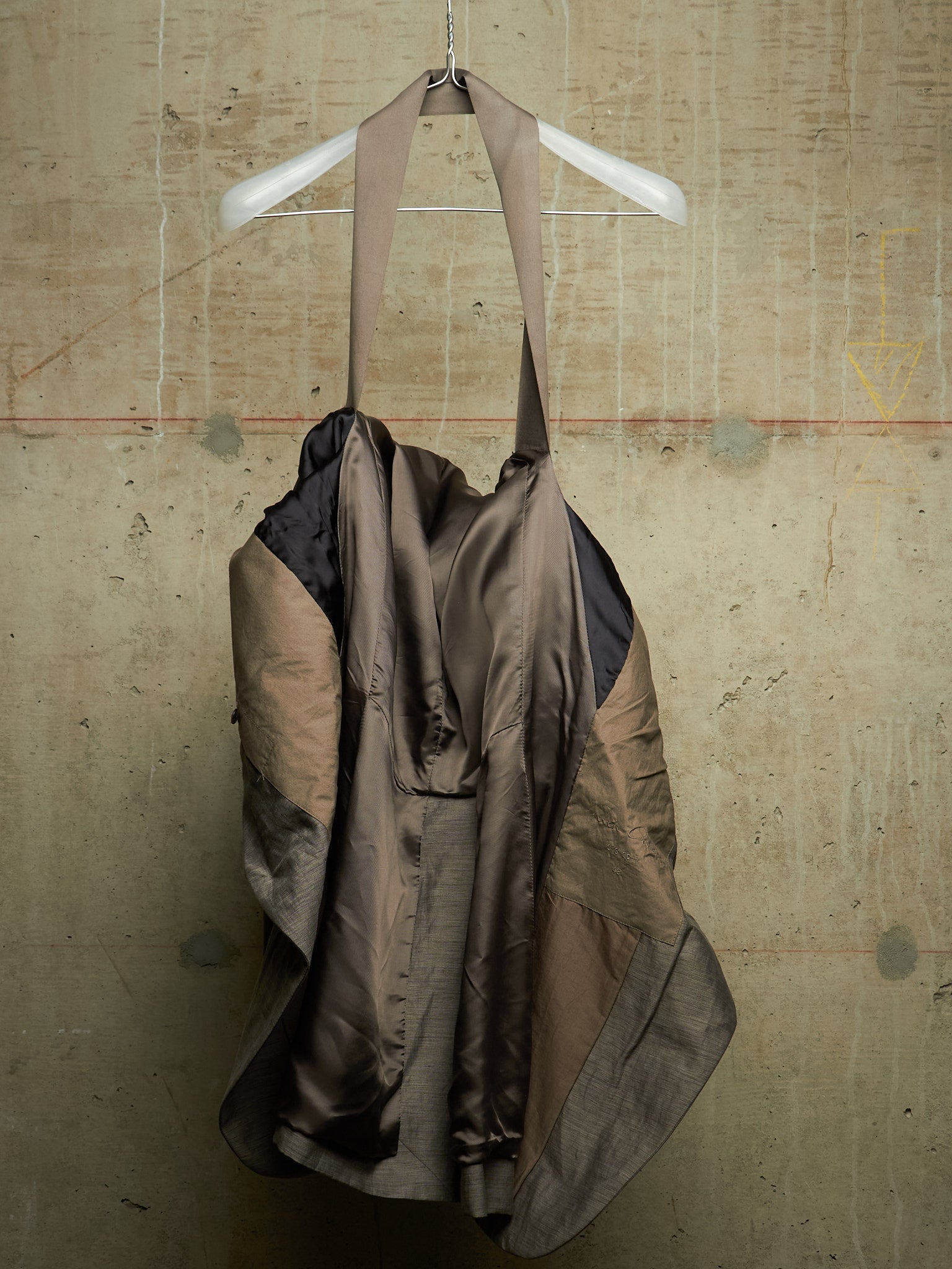 A draped, asymmetrical garment or dress made from layered taupe, brown, and black fabrics, hanging from a white plastic hanger against a textured concrete wall.