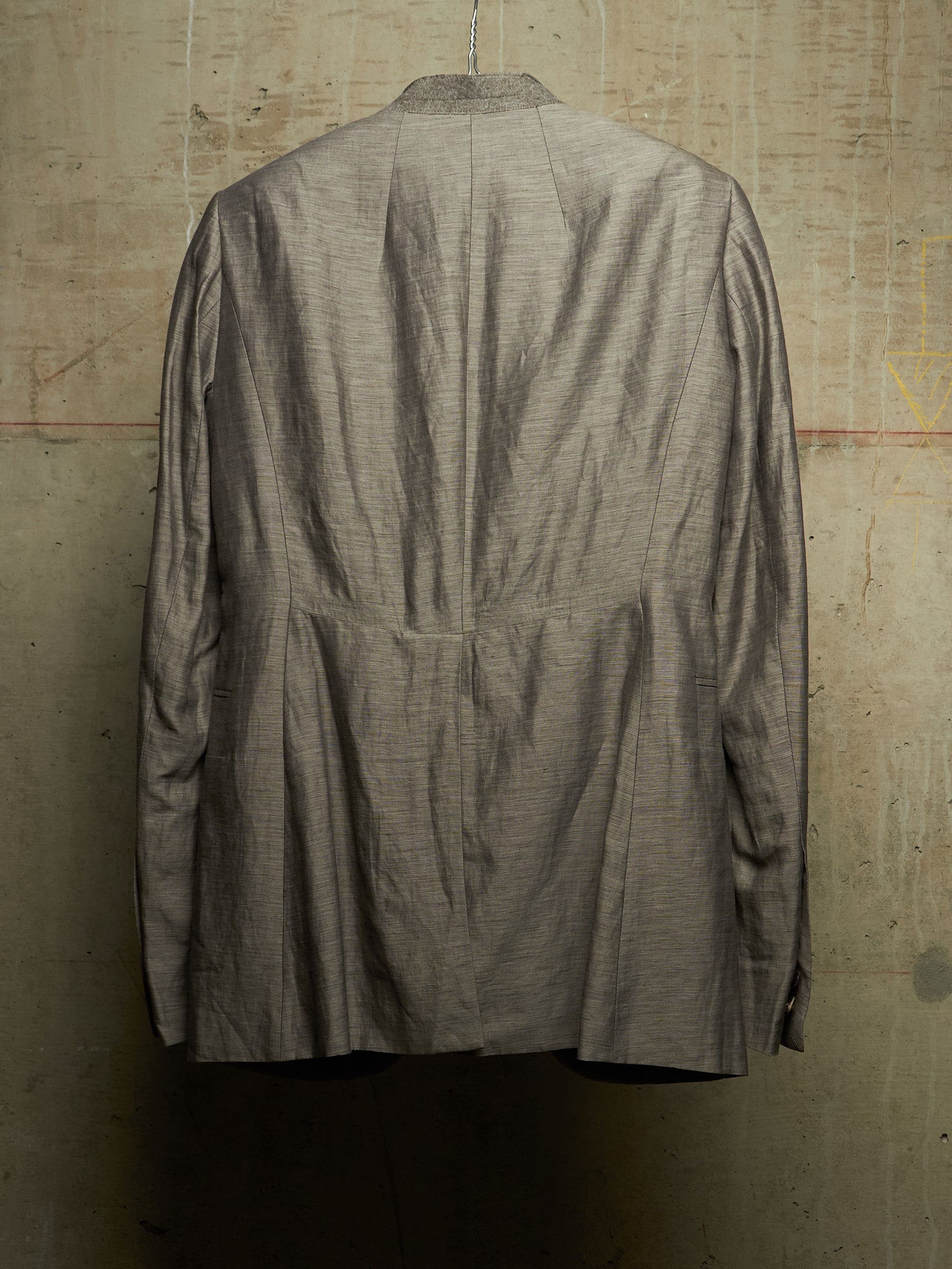 A textured gray linen-blend jacket hanging on a hanger against a rustic concrete wall, showing the back view with a central seam and pleated details.