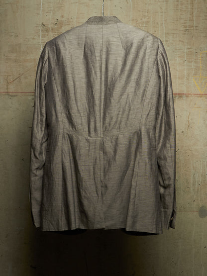 A textured gray linen-blend jacket hanging on a hanger against a rustic concrete wall, showing the back view with a central seam and pleated details.