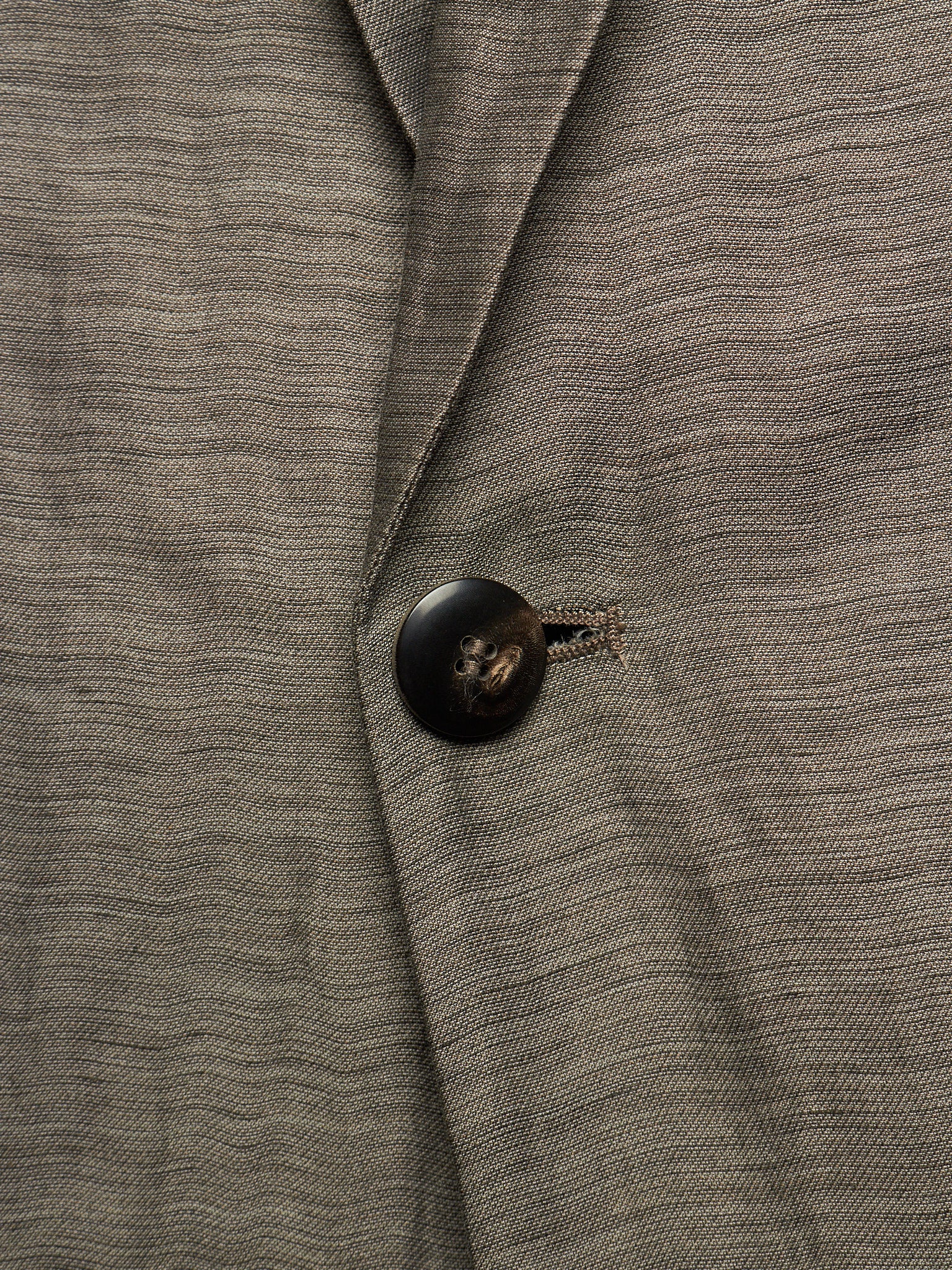 Close-up of a textured gray wool-blend blazer with a dark brown button and a small embroidered detail near the collar.