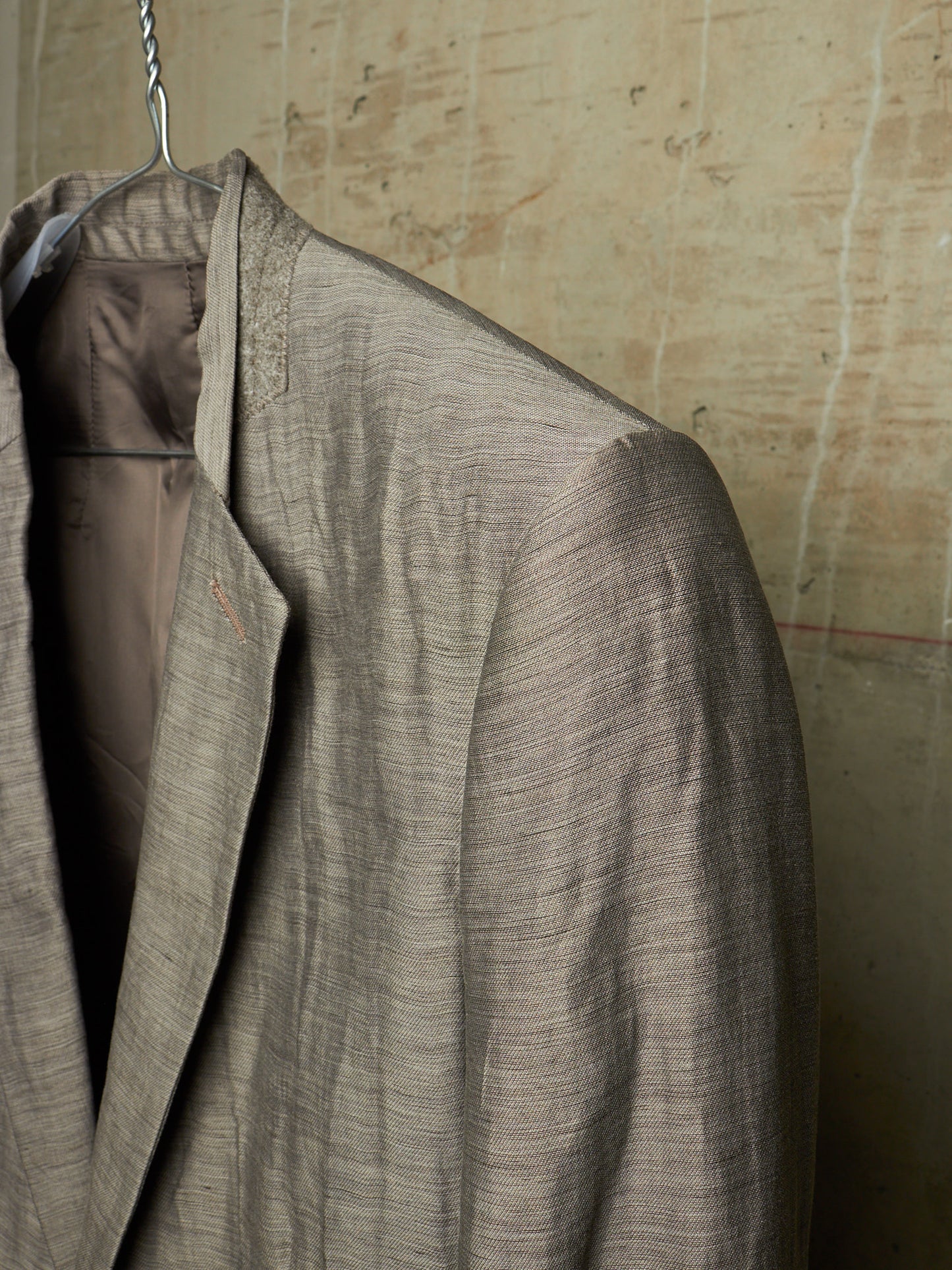 A close-up of a textured taupe-gray linen-blend blazer hanging on a metal hanger against a rustic, textured beige wall.