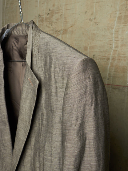 A close-up of a textured taupe-gray linen-blend blazer hanging on a metal hanger against a rustic, textured beige wall.