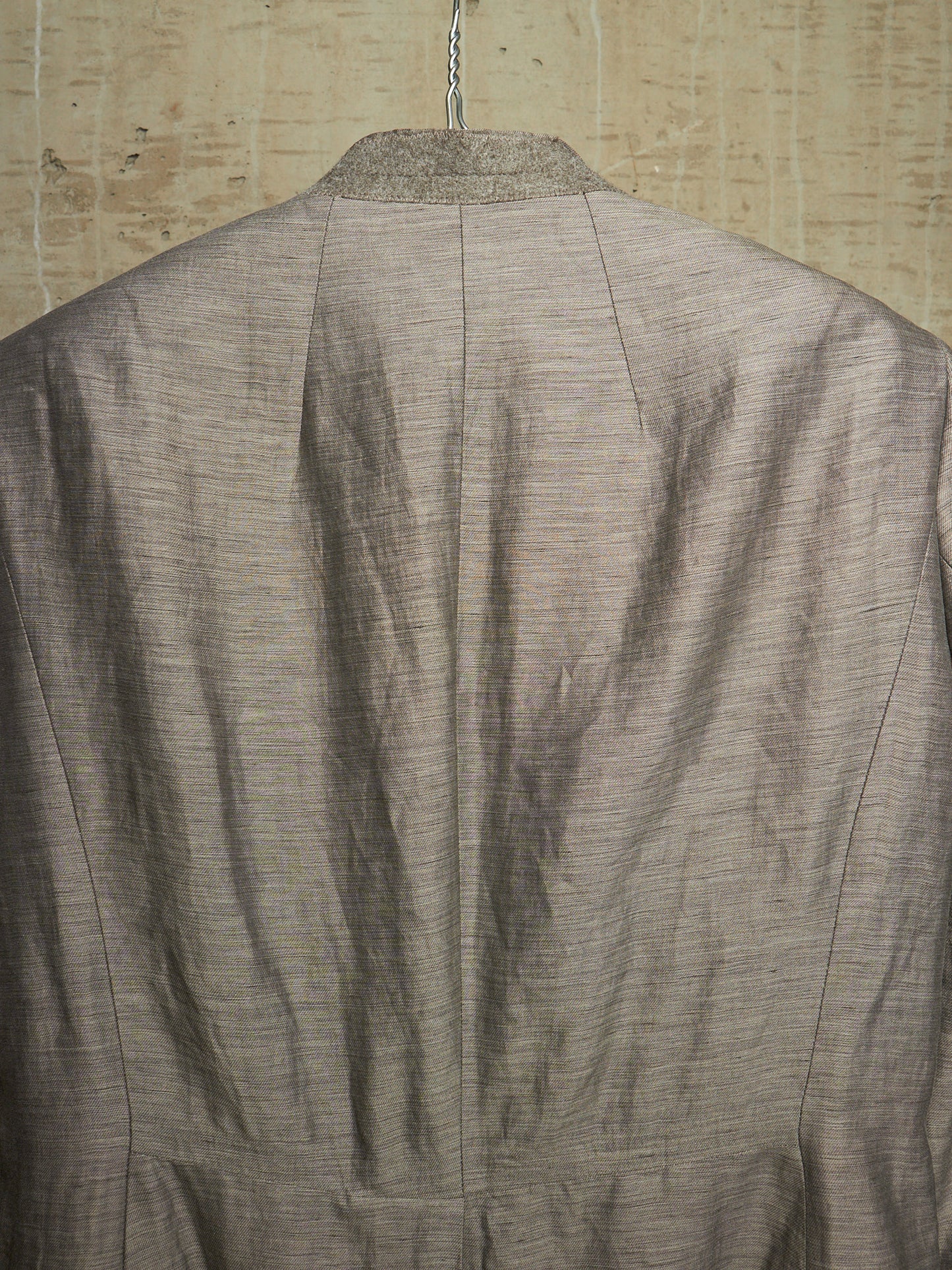 The back view of a textured taupe-gray linen jacket hanging against a rustic beige linen background.