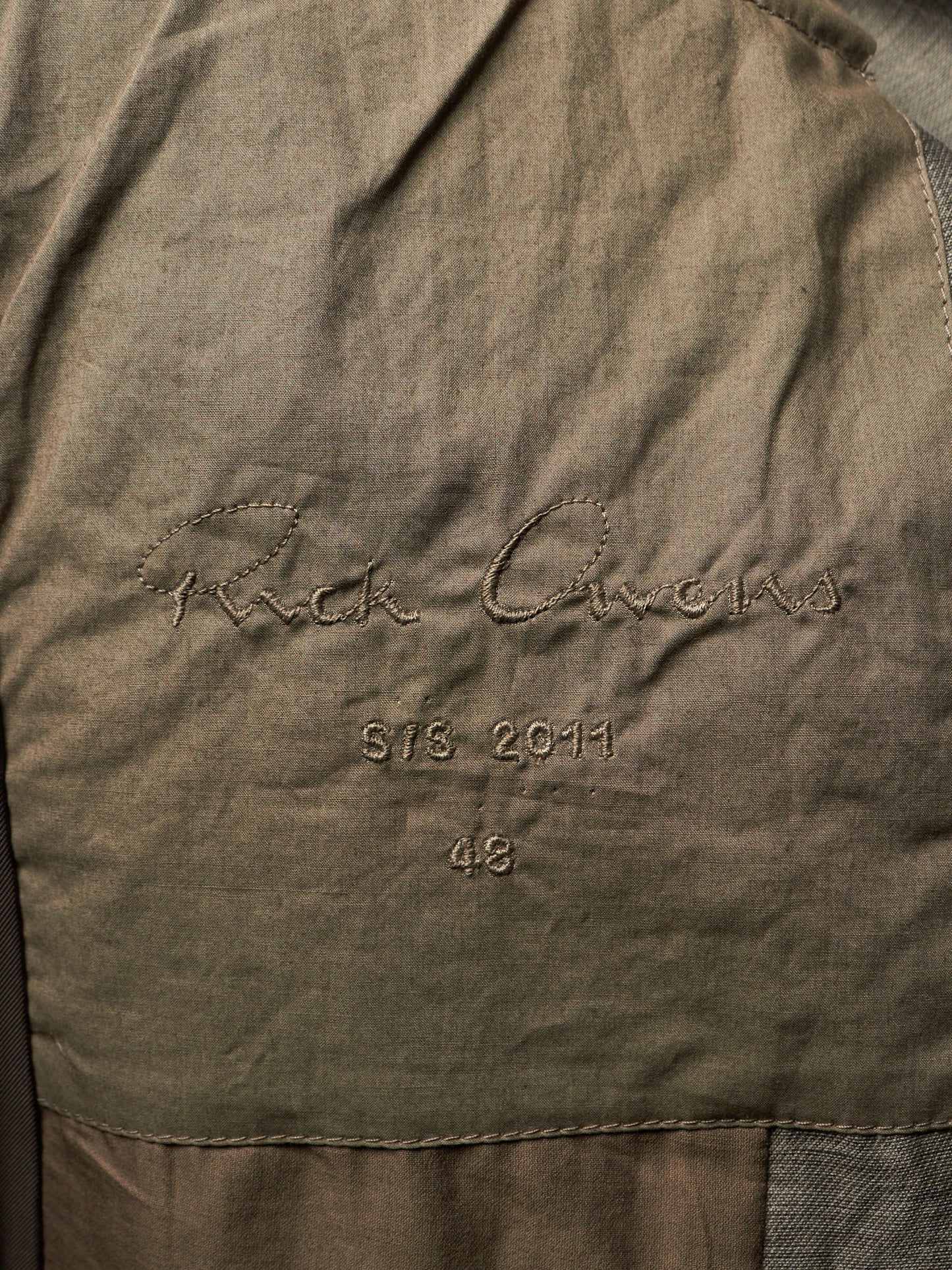 Close-up of a textured olive green fabric surface with embroidered script reading 'Rick Owens' and smaller stitched text 'STS. 2011' and '48'.
