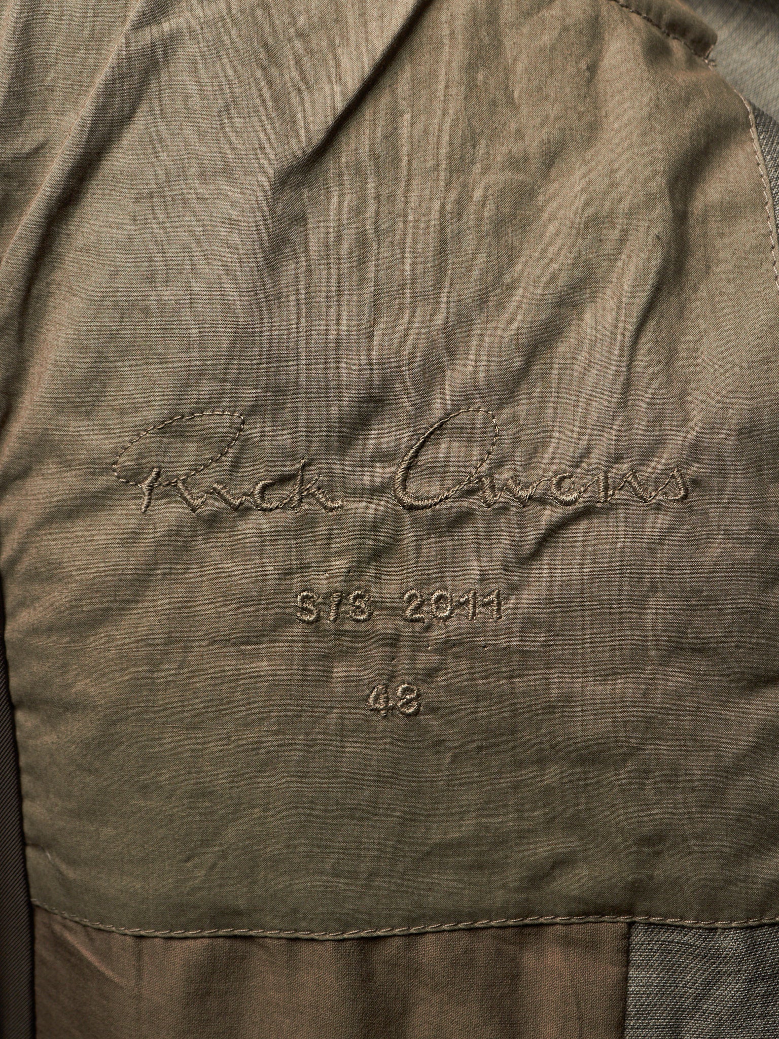 Close-up of a textured olive green fabric surface with embroidered script reading 'Rick Owens' and smaller stitched text 'STS. 2011' and '48'.