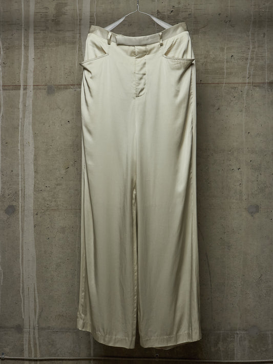 A pair of wide-leg trousers in a light cream or off-white color, hanging from a white plastic hanger against a textured concrete wall.