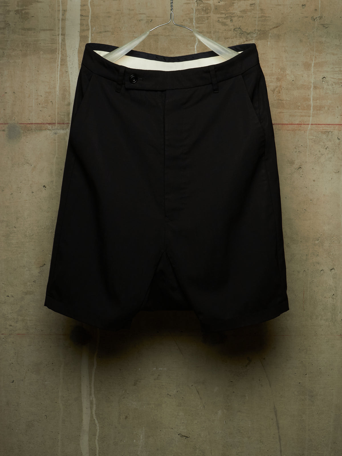 Black shorts hanging on a white hanger against a textured concrete wall.
