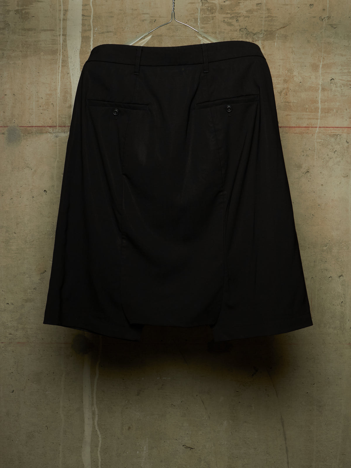 A black A-line skirt with two buttoned flap pockets, hanging on a clear plastic hanger against a textured concrete wall.