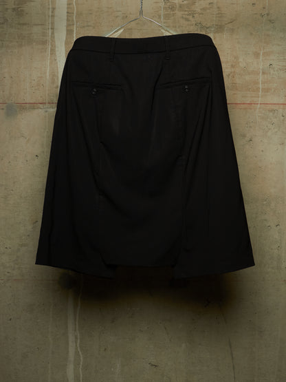 A black A-line skirt with two buttoned flap pockets, hanging on a clear plastic hanger against a textured concrete wall.