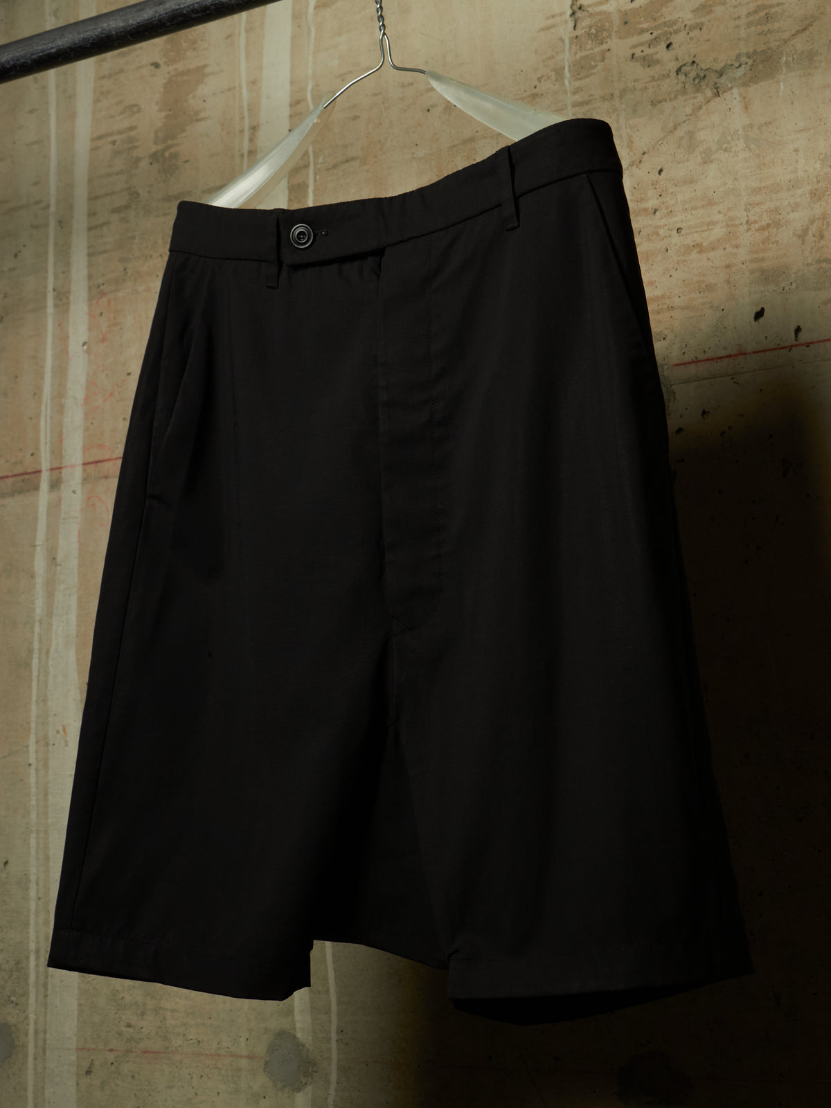 Black pleated shorts hanging on a metal hanger against a textured concrete wall.