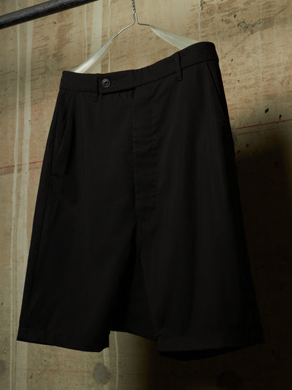 Black pleated shorts hanging on a metal hanger against a textured concrete wall.