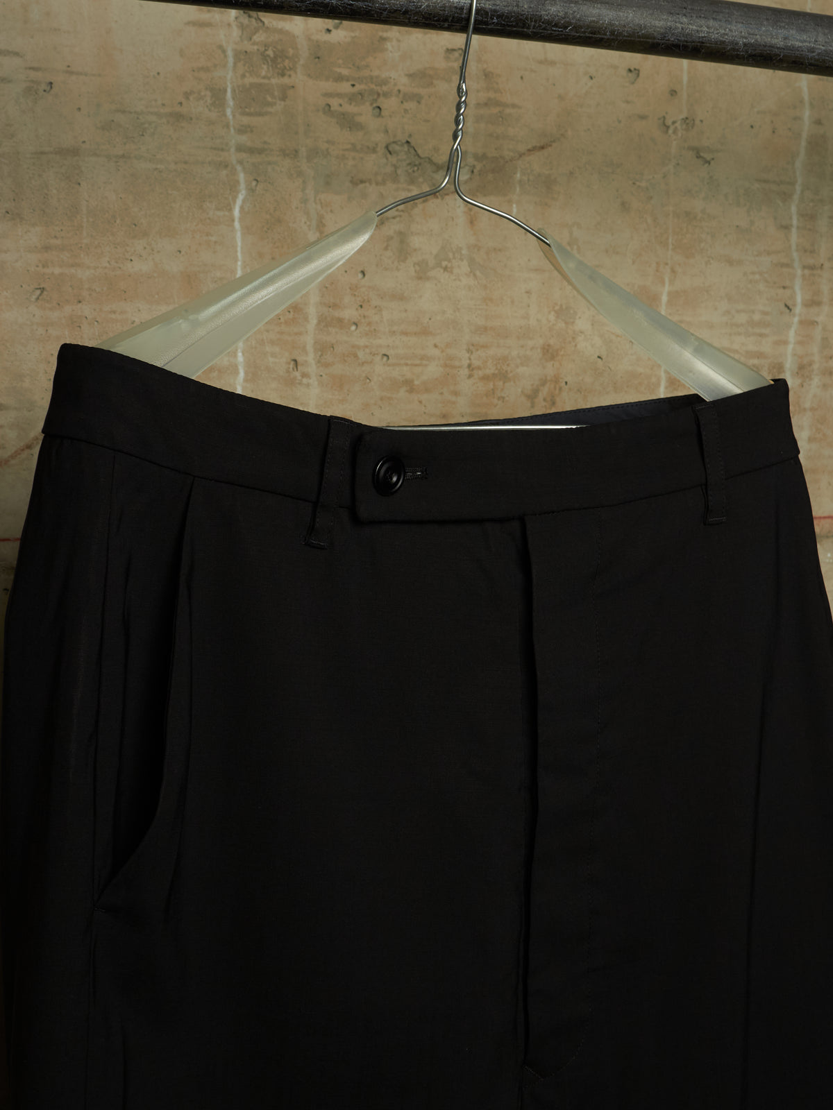 Black pleated trousers hanging on a clear plastic hanger against a textured beige wall.