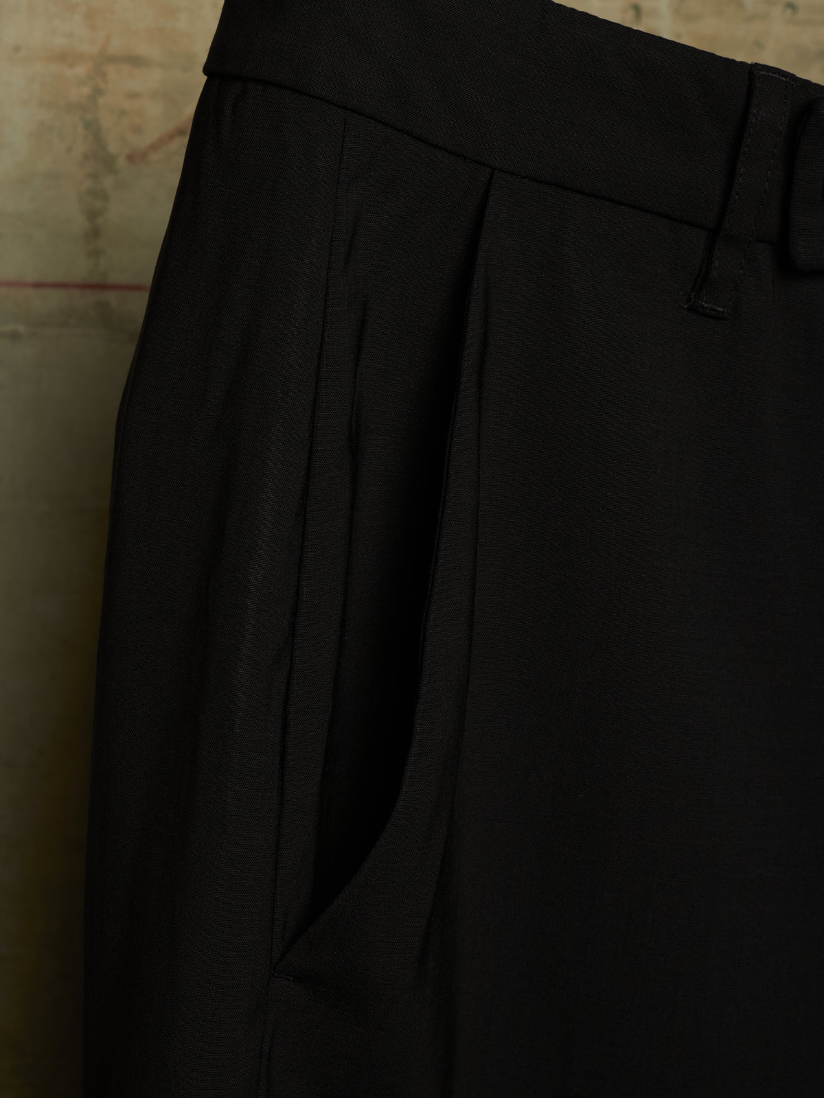 Close-up of black pleated trousers showing the waistband, belt loops, and side pocket against a textured beige background.