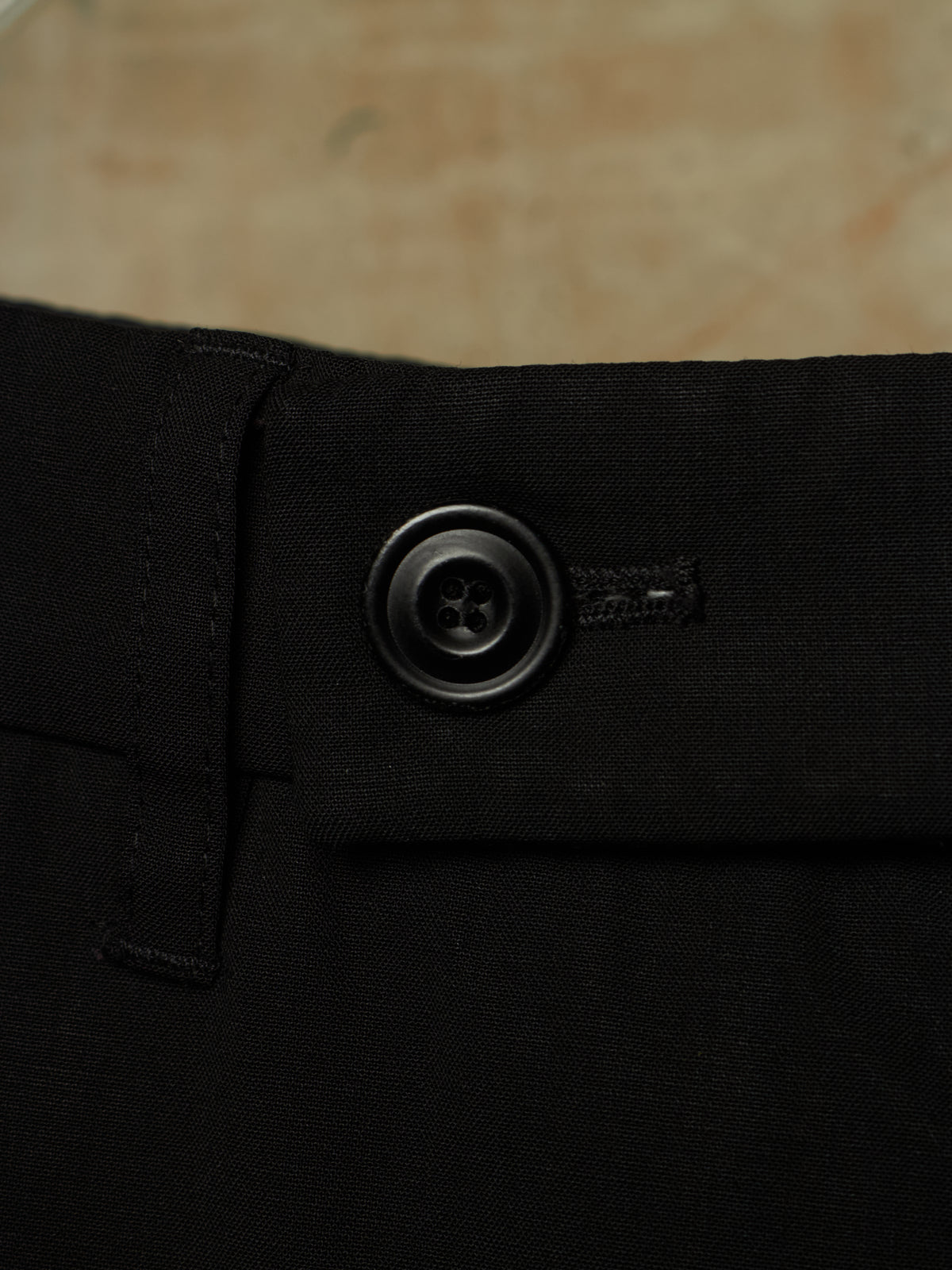 Close-up of black trousers showing the waistband, belt loop, and a dark metal button closure against a neutral beige background.