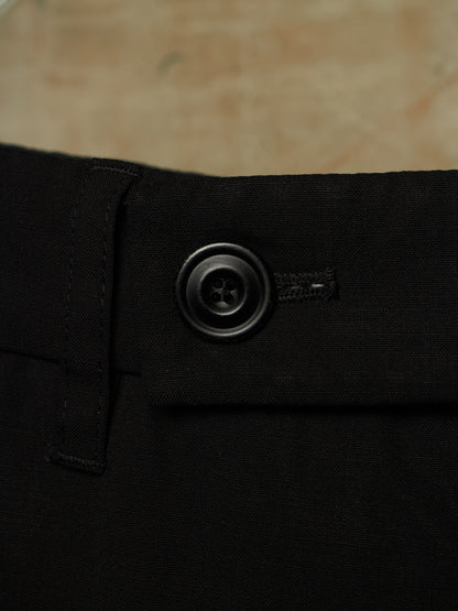 Close-up of black trousers showing the waistband, belt loop, and a dark metal button closure against a neutral beige background.
