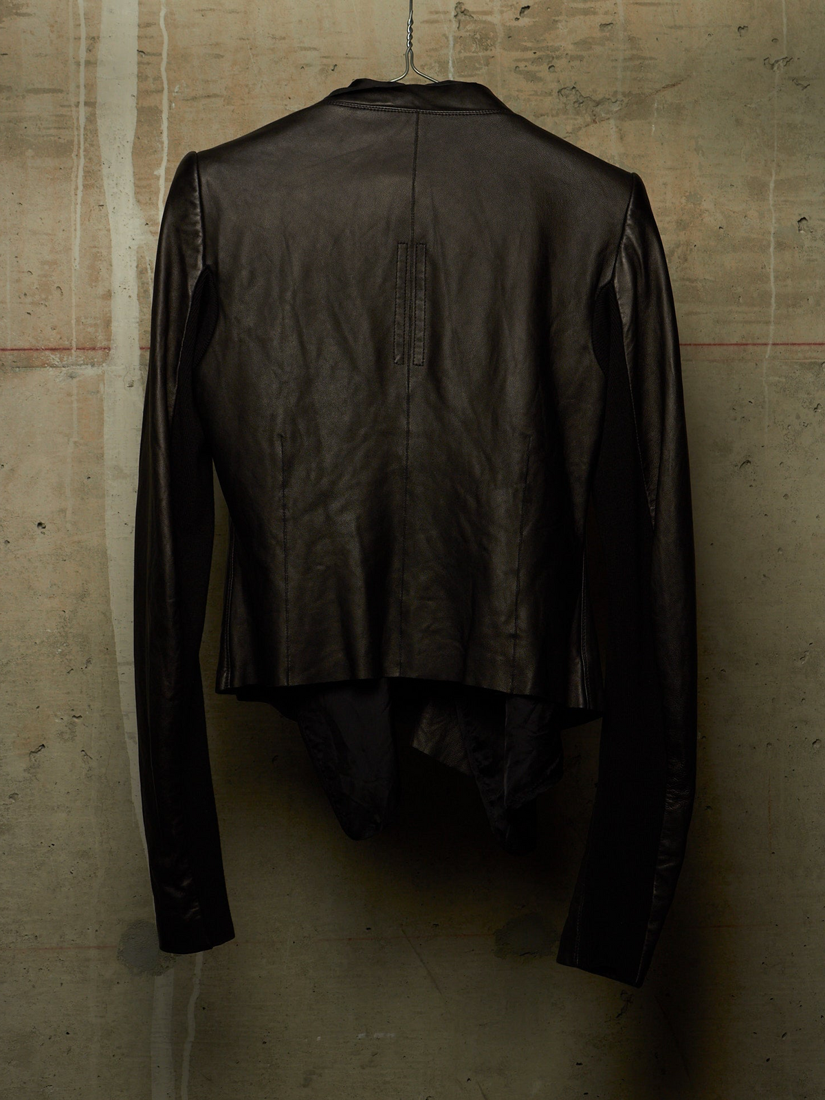 A black leather jacket with a unique asymmetrical, draped back design, hanging against a textured concrete wall.