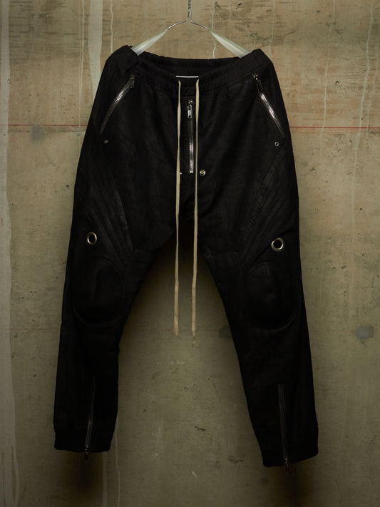 Black textured pants with silver zippers, metal grommets, and a cream-colored drawstring, hanging against a concrete wall.