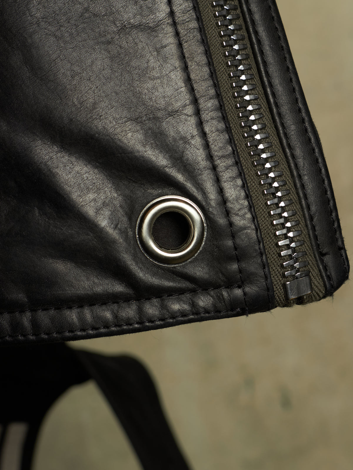 Close-up detail of a black leather jacket showing a silver metal grommet and a dark zipper with silver teeth.