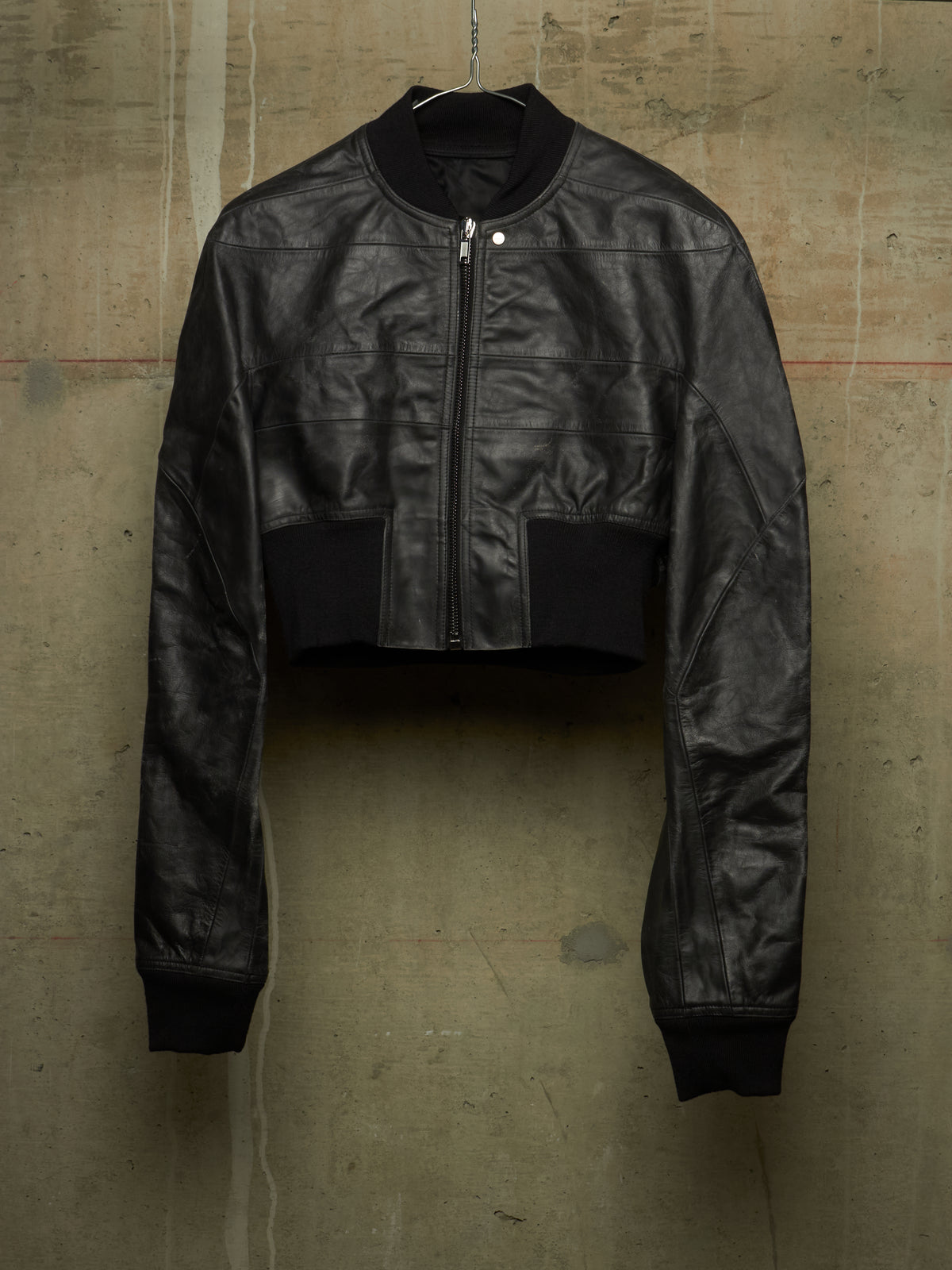 A black cropped leather jacket with a front zipper, ribbed collar, cuffs, and hem, hanging against a textured concrete wall.
