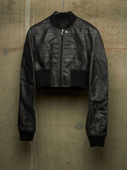 A black cropped leather jacket with a front zipper, ribbed collar, cuffs, and hem, hanging against a textured concrete wall.