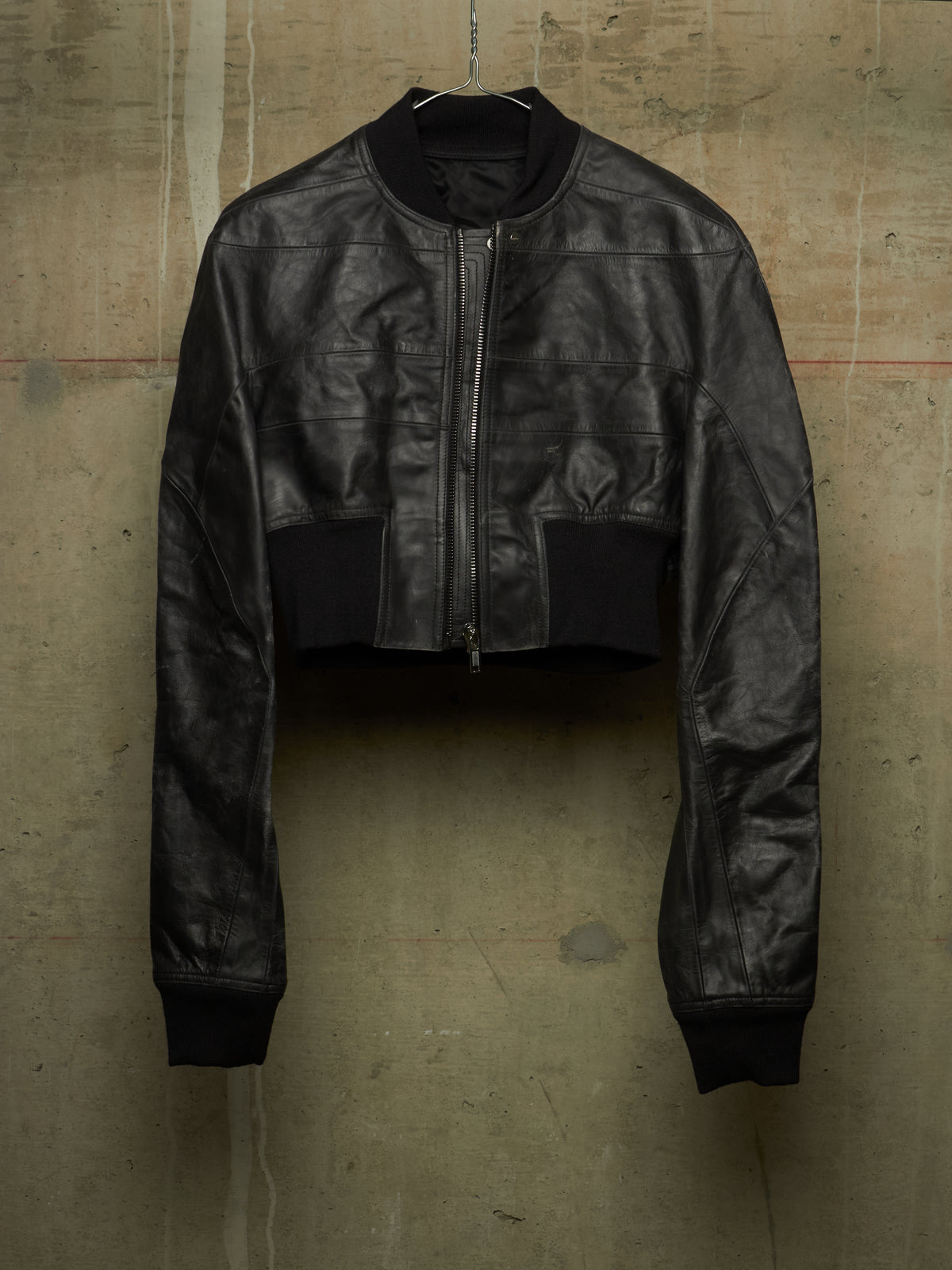 A black cropped leather jacket with a front zipper, ribbed collar, cuffs, and hem, hanging against a textured concrete wall.