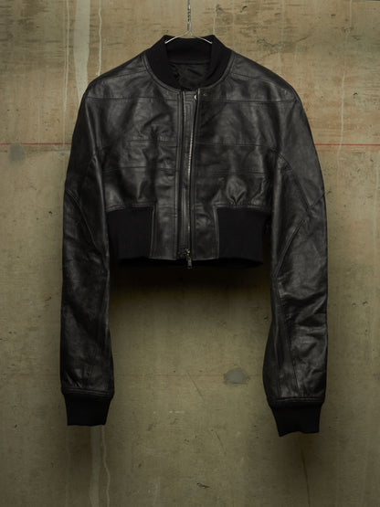 A black cropped leather jacket with a front zipper, ribbed collar, cuffs, and hem, hanging against a textured concrete wall.