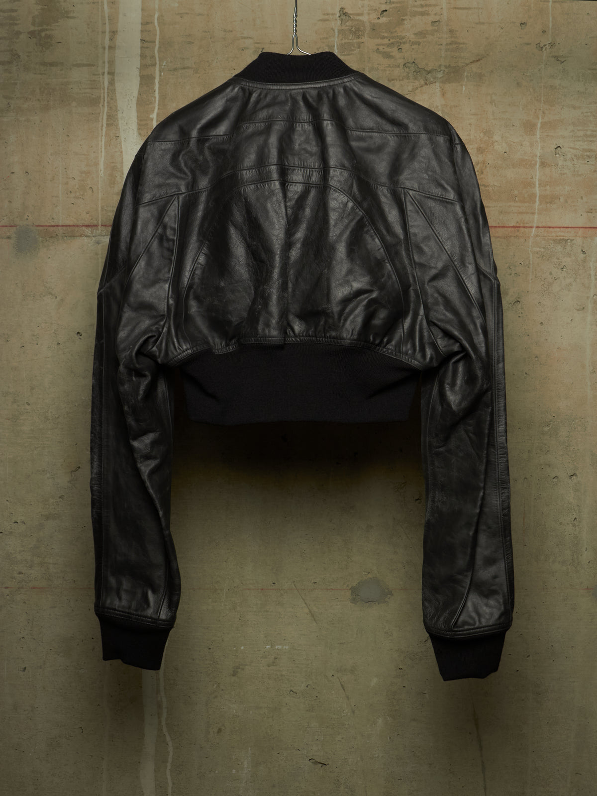 A cropped black leather jacket with long sleeves and ribbed cuffs, shown from the back hanging against a textured concrete wall.