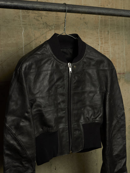 A black leather bomber jacket hanging on a metal hanger from a dark clothing rack against a textured concrete wall.