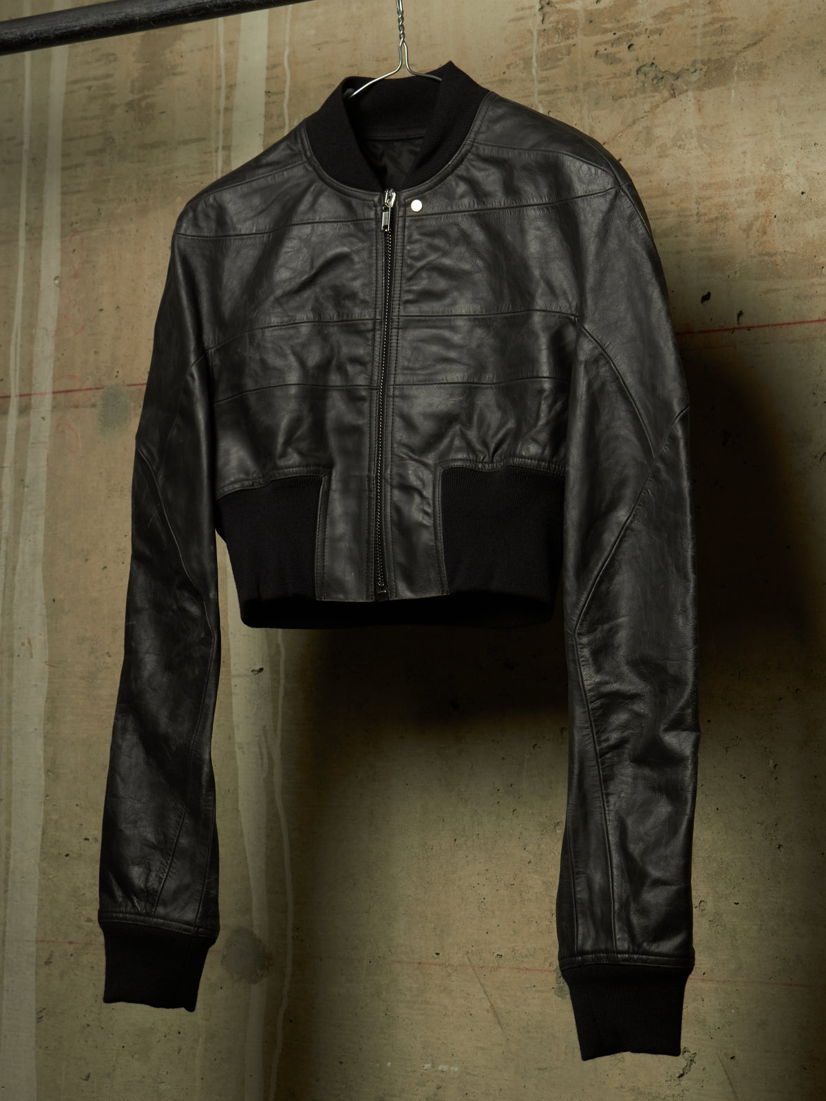 A black cropped leather jacket with a front zipper, ribbed collar, cuffs, and hem, hanging against a textured concrete wall.