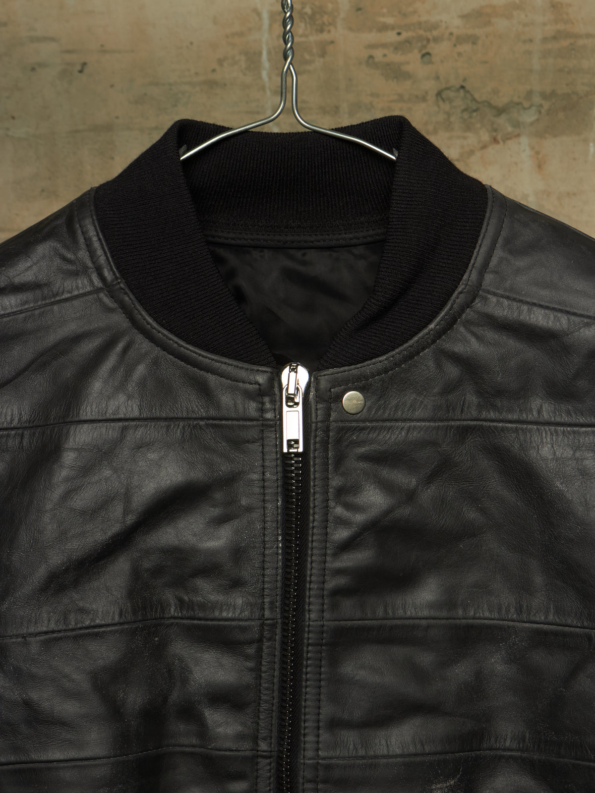 A close-up of a black leather jacket hanging on a metal hanger against a textured, warm-toned wall. The jacket features a black ribbed knit collar, a silver metal zipper, and a silver snap button.