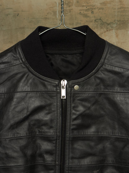 A close-up of a black leather jacket hanging on a metal hanger against a textured, warm-toned wall. The jacket features a black ribbed knit collar, a silver metal zipper, and a silver snap button.