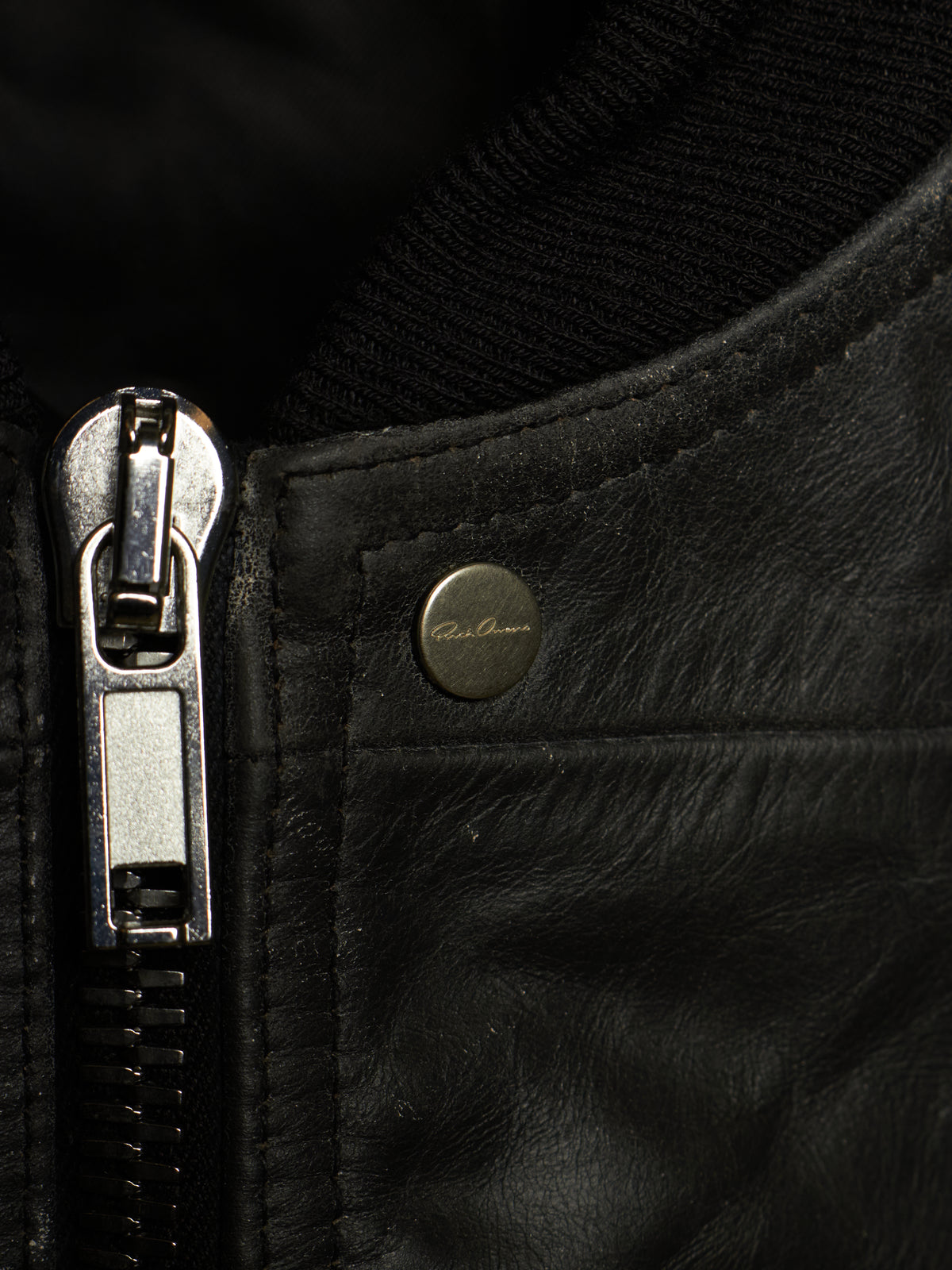 Close-up of a black leather jacket showing a silver metal zipper pull, a black ribbed knit collar, and a small round brass button with 'Paul Reed' branding.