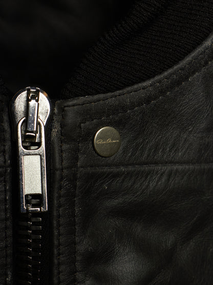 Close-up of a black leather jacket showing a silver metal zipper pull, a black ribbed knit collar, and a small round brass button with 'Paul Reed' branding.