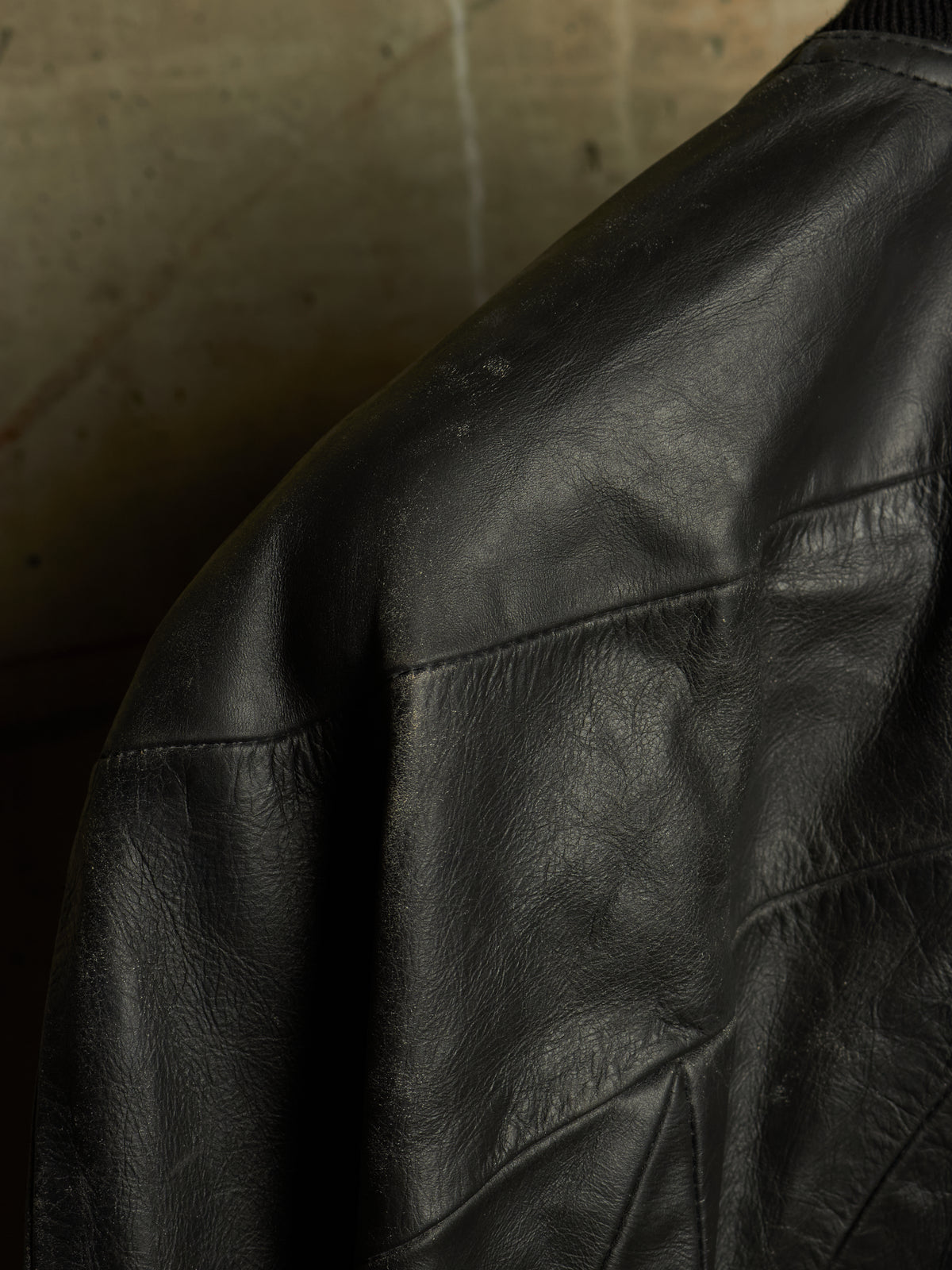 Close-up of a black leather jacket showing the texture and stitching on the shoulder and upper sleeve, set against a dark concrete background.